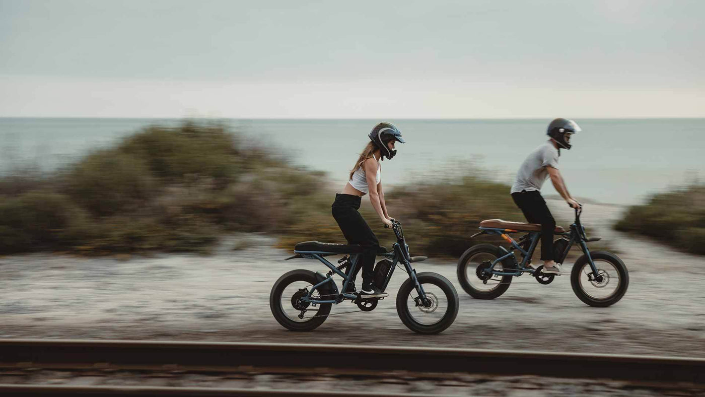 Two young riders on Super73 ebikes cruising along a trail near the beach with helmets on